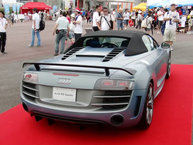 The image shows a grey Audi R8 Spyder parked on a red carpet in front of a crowd of people, some of...