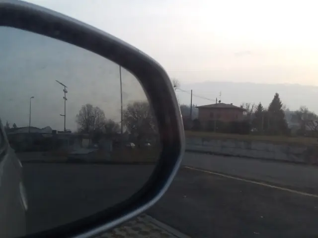 The image shows a car's side view mirror on the side of a road, with the reflection of a building,...