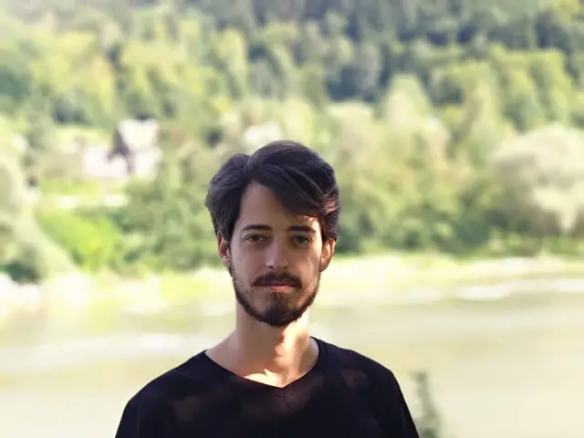 The image shows a man standing in front of a lake, wearing a black t-shirt and jeans. The lake is...