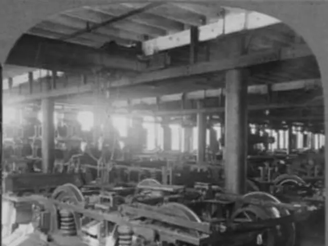 The image shows a black and white photo of an electric motor and car truck in a factory. The...