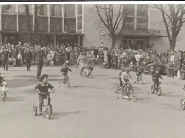 The image shows a group of children riding bicycles down a street, with a crowd of people standing...