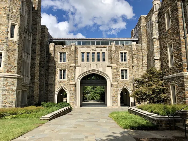 The image shows a large stone building with a walkway in front of it, surrounded by lush green...