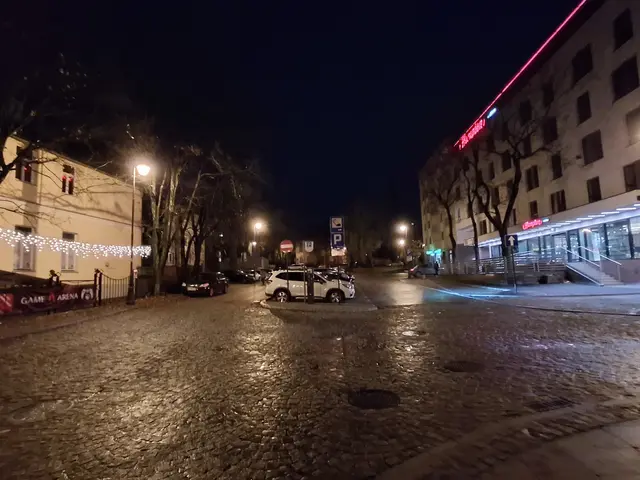 The image shows a city street at night with cars parked on the side of it. There are street lights...