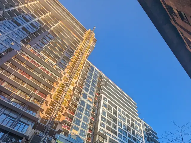 The image shows a tall building under construction in the middle of a city, with vehicles on the...